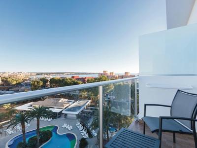 Balcon d'hôtel avec chaises, vue sur piscine et paysage urbain au loin sous un ciel bleu clair.