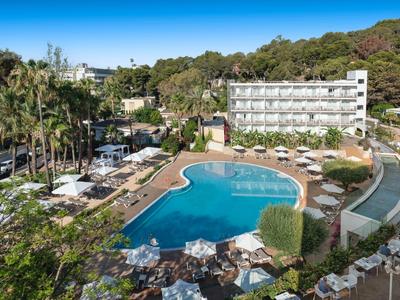 View of a pool with sun loungers and a multi-story hotel in the background in a green setting.