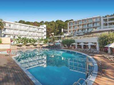 Hotel con piscina all'aperto, sedie a sdraio e ombrelloni davanti a edifici bianchi sotto un cielo azzurro