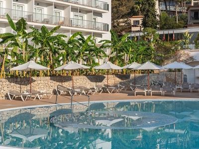 Hotel pool with sun loungers and umbrellas in front of a building and tropical plants.