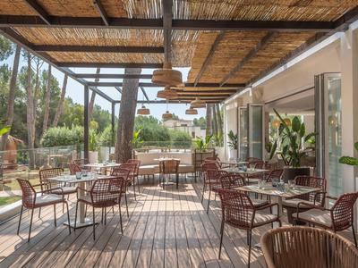 Covered terrace with tables and chairs, surrounded by palm trees and green plants.