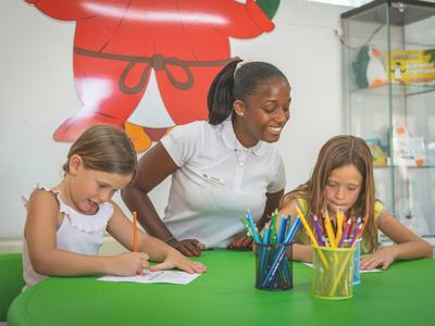 Deux filles et une animatrice sont assises à une table verte en train de dessiner avec des crayons de couleur.