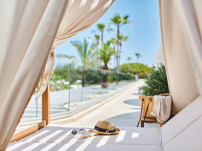 Výhled skrz bílé závěsy na bazénovou oblast s palmami a pohodlným posezením.
