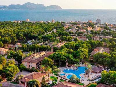 View of a resort with a pool, surrounded by trees, near the sea and distant mountains.