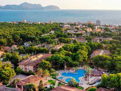 Pohled na hotelový komplex s bazénem, obklopený zelení s výhledem na moře a hory.