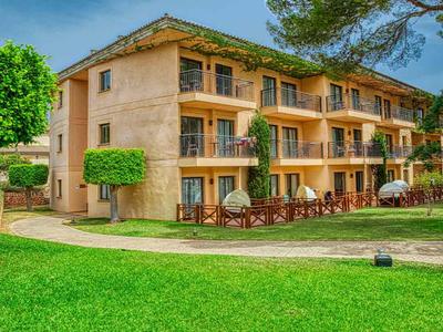 Třípatrový hotel s balkónovými pokoji, obklopený zeleným trávníkem a stromy pod modrou oblohou.