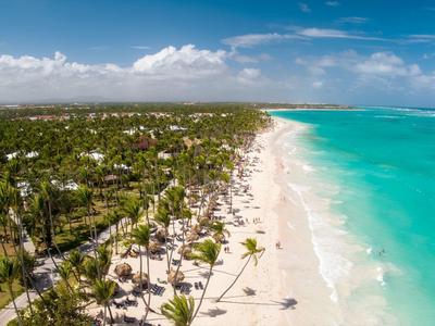 Langer Sandstrand mit türkisfarbenem Wasser und vielen Palmen unter blauem Himmel mit Wolken.