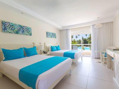 Bright hotel room with two double beds, turquoise accents, and balcony with palm tree view.