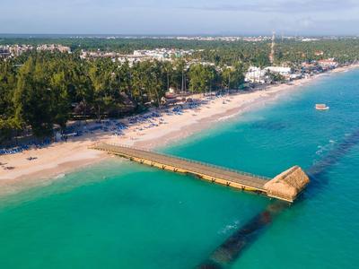 Luftaufnahme eines langen Stegs, der über türkisfarbenes Wasser zu einem Sandstrand mit Bäumen führt.