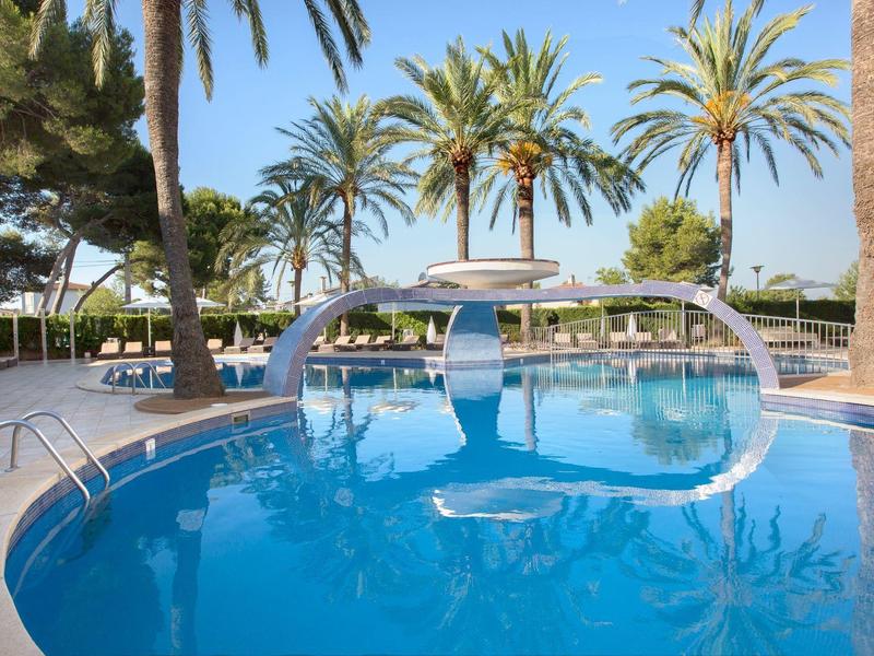 Swimming pool with sunshades and palm trees in a tropical hotel area under clear sky.
