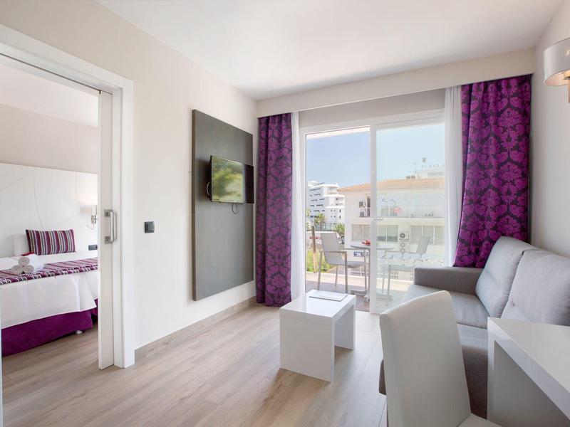 Modern hotel room with balcony, seating area, TV, and adjoining bedroom.