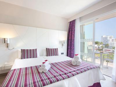 Habitación de hotel luminosa con cama doble, ropa de cama morada y balcón con vistas.