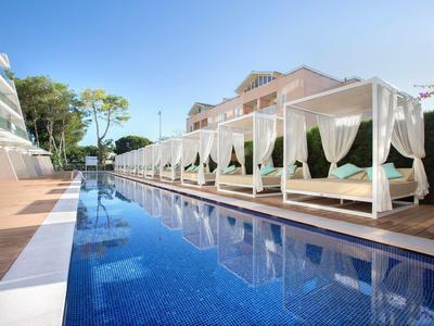 Piscina exterior moderna con camas balinesas y cortinas blancas en un hotel bajo cielo despejado.