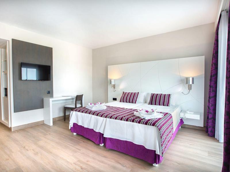 Habitación de hotel moderna con cama doble, paredes claras, suelo de madera y detalles morados.