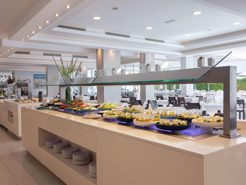 Área de buffet con varias comidas y mesas blancas modernas en un restaurante luminoso.