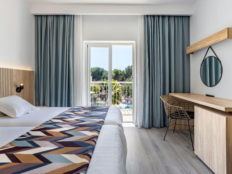 Lichte hotelkamer met bed, houten meubels en balkon met uitzicht op groen.