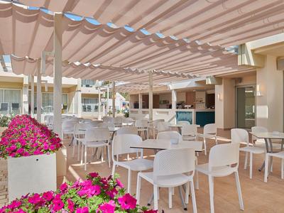 Leerer Außenbereich eines Cafés mit weißen Stühlen und rosa Blumen unter einem Sonnendach.