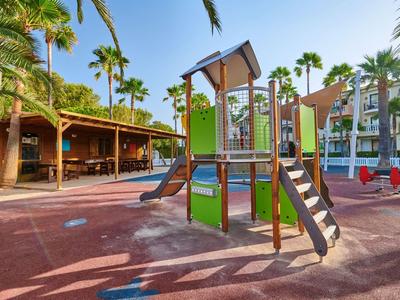 Aire de jeux pour enfants avec structure d'escalade et toboggan près d'un espace couvert et des palmiers.