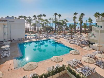 Zone de piscine avec chaises longues blanches et parasols à côté des bâtiments de l'hôtel et des palmiers.