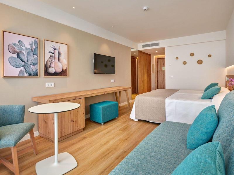 Chambre d'hôtel lumineuse avec deux lits, canapé bleu, fauteuil et décorations modernes.