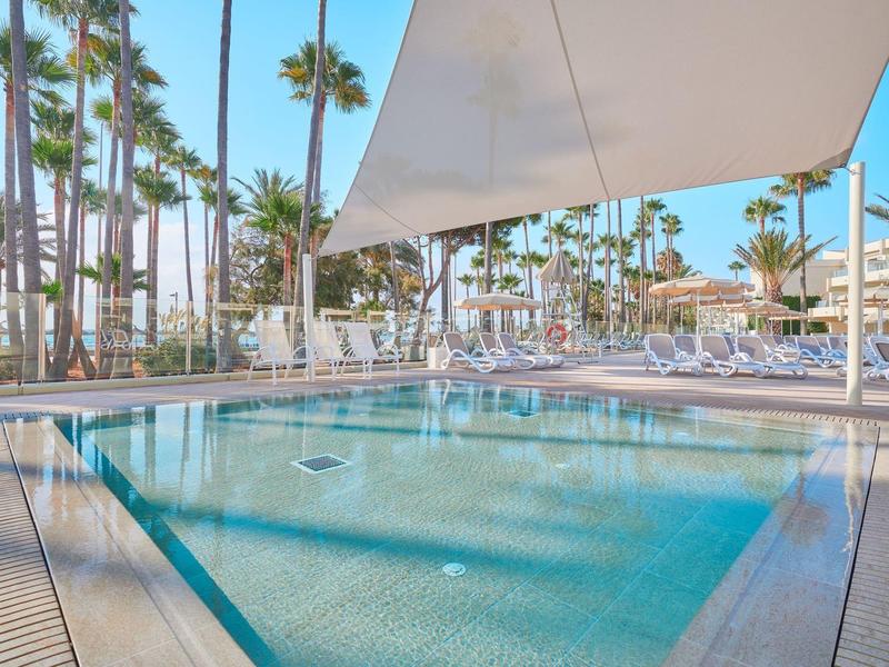 Piscine d'hôtel claire avec palmiers et voile d'ombrage par temps ensoleillé.
