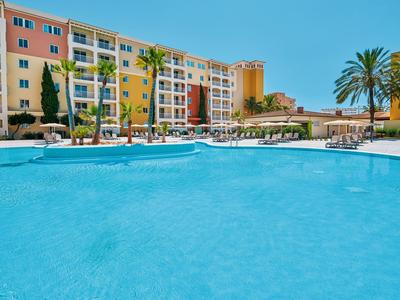 Gran piscina al aire libre con tumbonas frente a un edificio de hotel de varios pisos bajo un cielo azul claro.