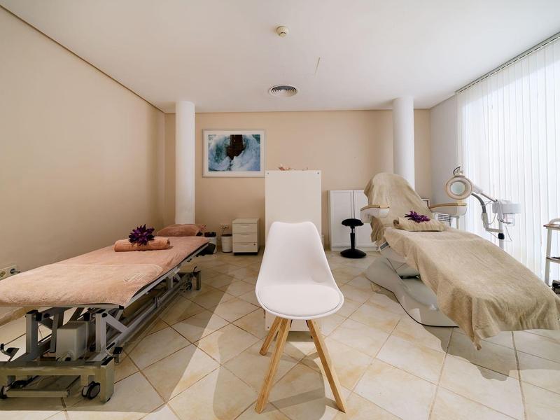 Helles Behandlungszimmer mit zwei Behandlungsbetten, Stühlen und natürlichem Licht durch große Fenster.