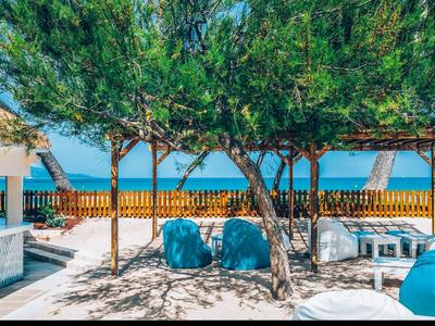 Sunny terrace with cushions, trees, and sea view behind a wooden fence.