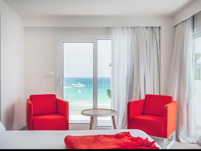 Bright hotel room with red armchairs, small table, and sea view.