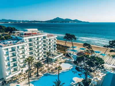 Hotel con grande piscina accanto a una spiaggia sabbiosa e al mare con colline sullo sfondo.