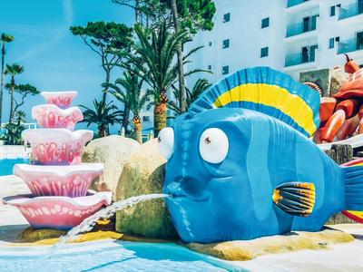 Fontana a forma di pesce colorato accanto a una piscina con palme e hotel sullo sfondo.