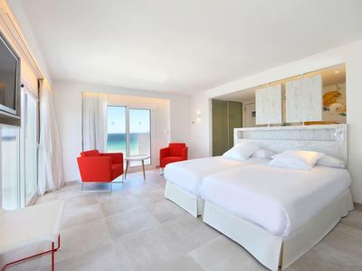 Camera d'hotel luminosa con due letti e due poltrone rosse vicino alla finestra.