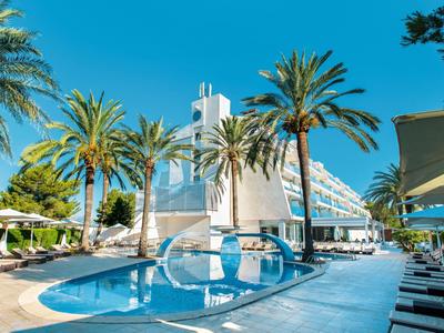 Nowoczesny kompleks hotelowy z basenem, palmami i leżakami pod jasnym niebieskim niebem.