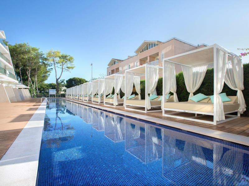 Espace piscine moderne avec lits blancs et rideaux, entouré d'eau bleue claire et d'arbres.