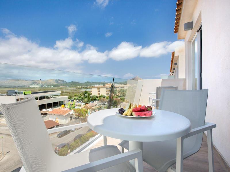 Balcon avec table blanche, chaises, corbeille de fruits, vue sur ville et collines.