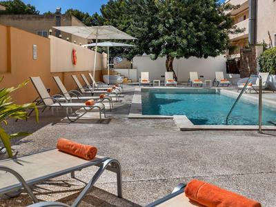 Hotel pool with lounge chairs, umbrellas, and a tree in the background on a sunny day.