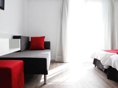 Bright room with simple bed, black bench, and red stool by a window with curtains.