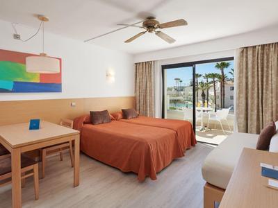 Hotel room with double bed, table, chairs, ceiling fan, and terrace overlooking palm trees.