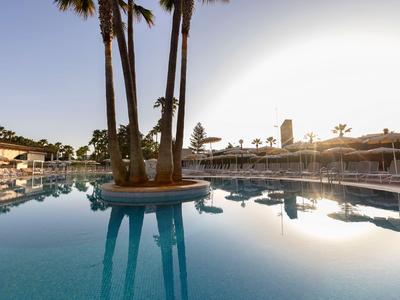 Gran piscina de hotel con isla de palmeras y luz solar reflejada al atardecer.