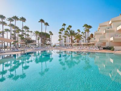 Gran piscina de hotel con palmeras y tumbonas bajo un cielo azul despejado