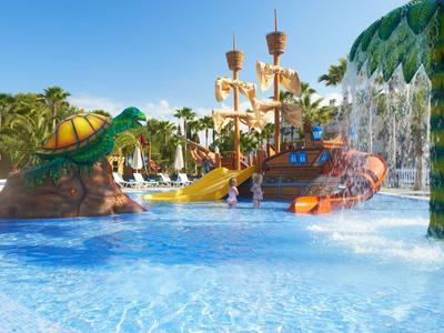 Colorido parque acuático con barco pirata, tortuga y palmeras en una piscina soleada.