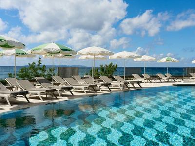 Grande piscina con lettini e ombrelloni vicino al mare sotto un cielo azzurro.