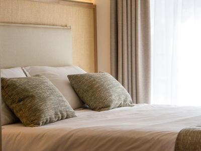 Gemütliches Bett mit weißen Laken, zwei grünen Kissen und hellem Vorhang im Hintergrund.