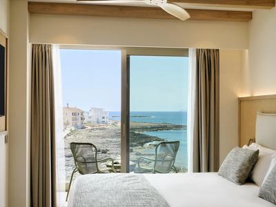 Helles Schlafzimmer mit weißer Bettwäsche und Balkon mit Stühlen und Meerblick im Hintergrund.