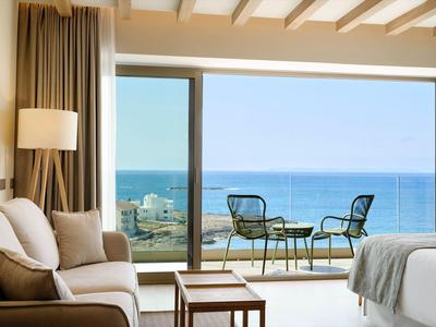 Helles Zimmer mit Sofa, Tisch, Bett und Balkon mit Blick auf das blaue Meer unter klarem Himmel.