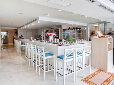 Area bar d'hotel luminosa con bancone bianco e sgabelli blu accanto a buffet e bevande.