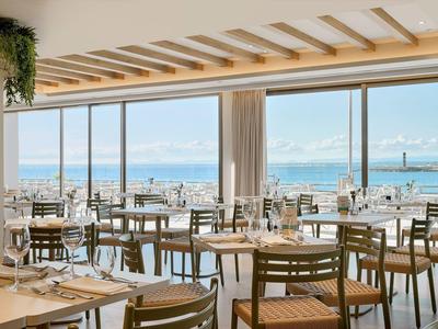 Modern restaurant with wooden chairs and tables overlooking the sea through large windows.
