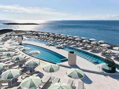 Área moderna de piscina junto al mar con tumbonas y sombrillas bajo un cielo azul.