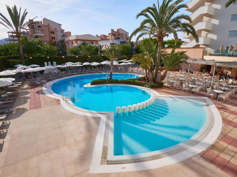 Hotel pool with palm trees, umbrellas, and chairs next to a restaurant area.