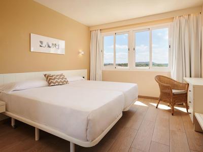 Bright hotel room with large bed, wicker chair, and window showing blue sky outside.